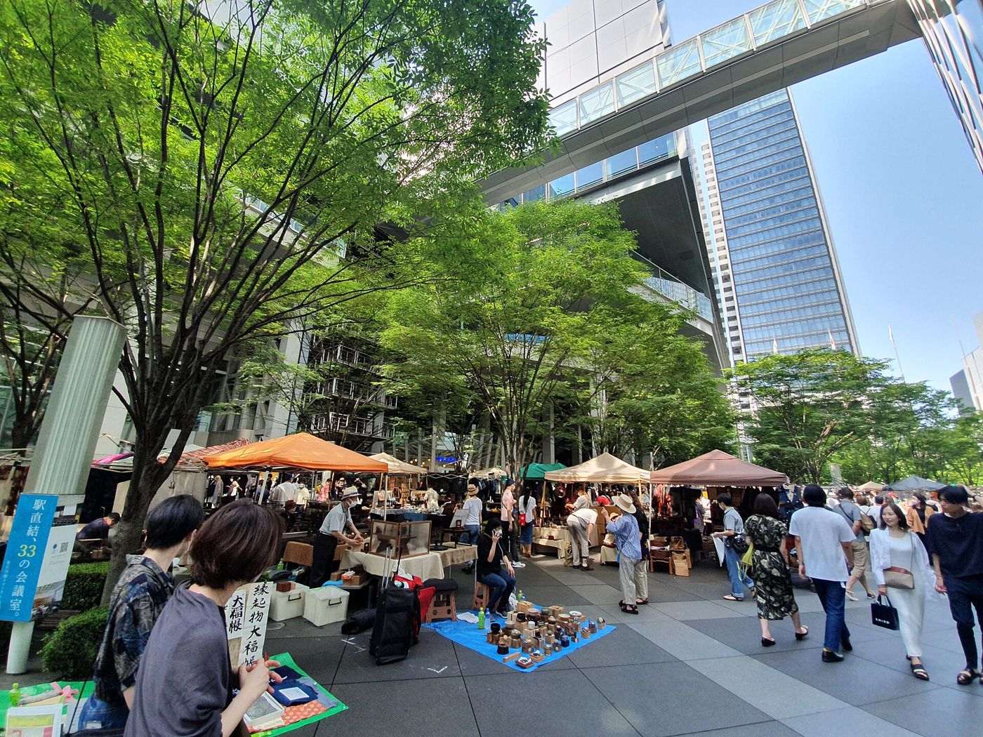Stalls of a weekend flea market under trees between glass buildings, busy with shoppers